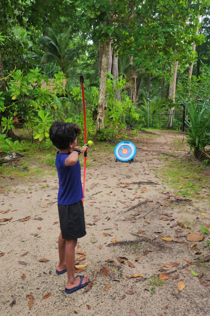 child in blue shirt and black shorts aiming a his bow and arrow at the archery board in the distance. the archery activity at taj havelock is set amongst a small forest of trees.
