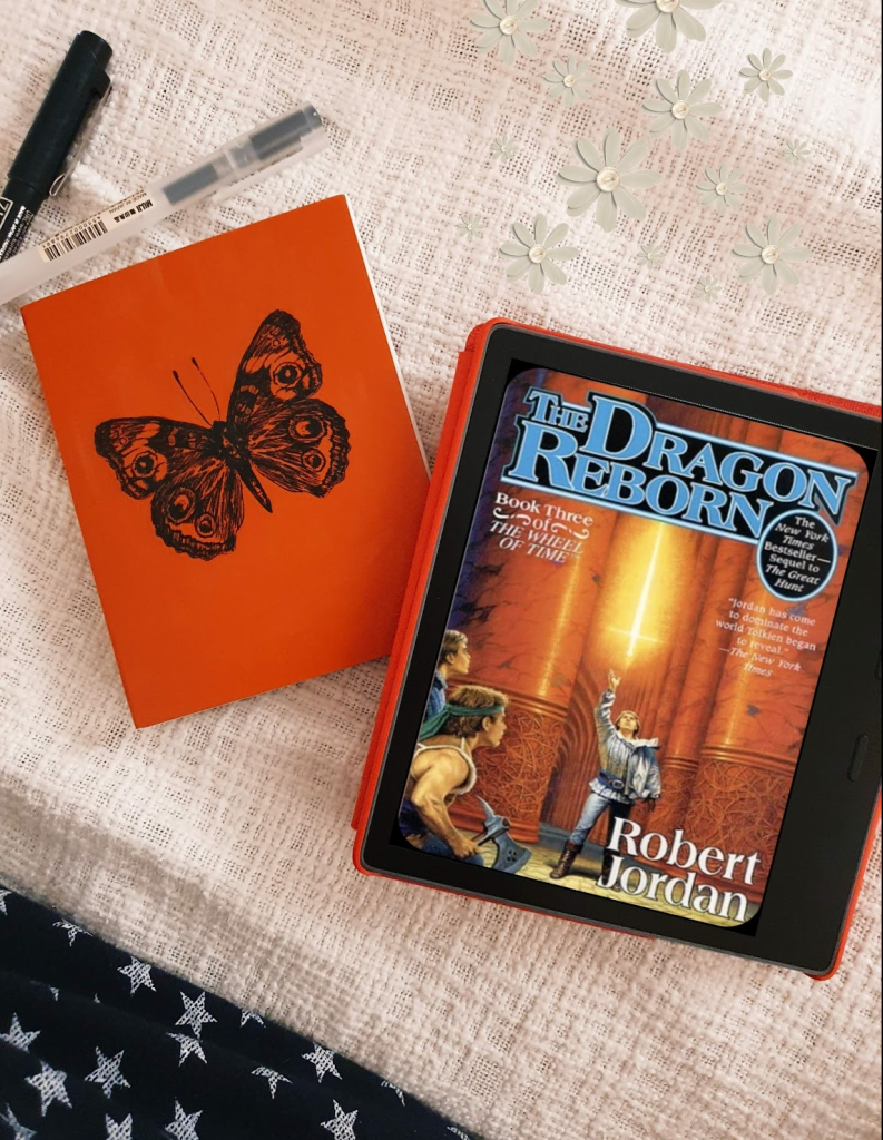 Robert Jordan's Wheel of Time Book 3 the dragon reborn cover on a Kindle Oasis with a notebook in red with a butterfly cover and 2 pens on a white bedcover